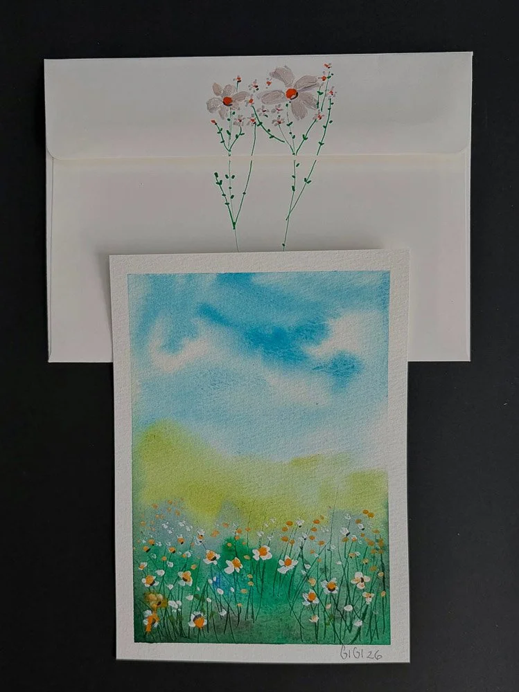 A watercolor painting of a field of white and orange daisies under a blue sky with some clouds; the painting is placed on top of an envelope with a drawing of flowers on it.