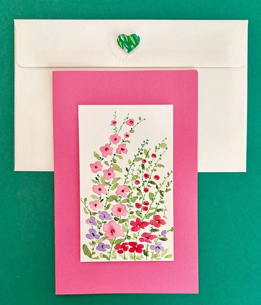 A floral greeting card with pink, purple, and red flowers on green stems, placed on a pink textured background, with a white envelope with a green heart sticker on top of it.
