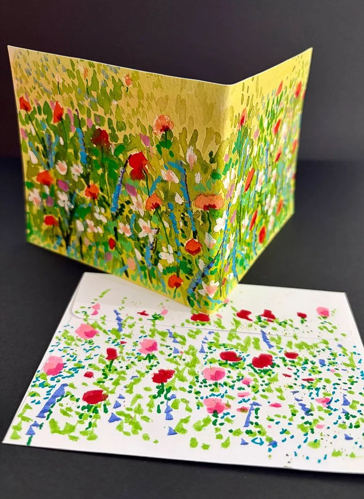 Colorful hand-painted greeting card with floral pattern on a black background, with an unfolded card showing a garden scene and a fallen floral design on a white sheet.