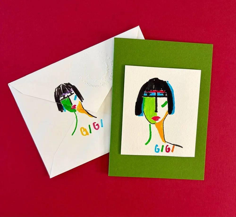A colorful, abstract portrait of a woman with black hair, green face, red lips, and various colored lines, on a white card and an envelope, set on a red background. The card has the name 'Gigi' written in multicolored letters.