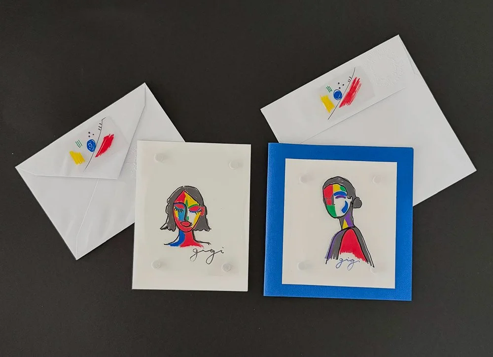 Two handwritten greeting cards with colorful abstract portraits, each signed 'gigi,' and two white envelopes with matching abstract designs on the flaps, all placed on a black surface.