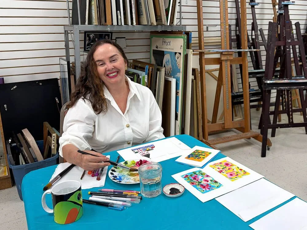 Woman sitting at a table with art supplies, smiling, with colorful paintings and brushes, in an art studio.