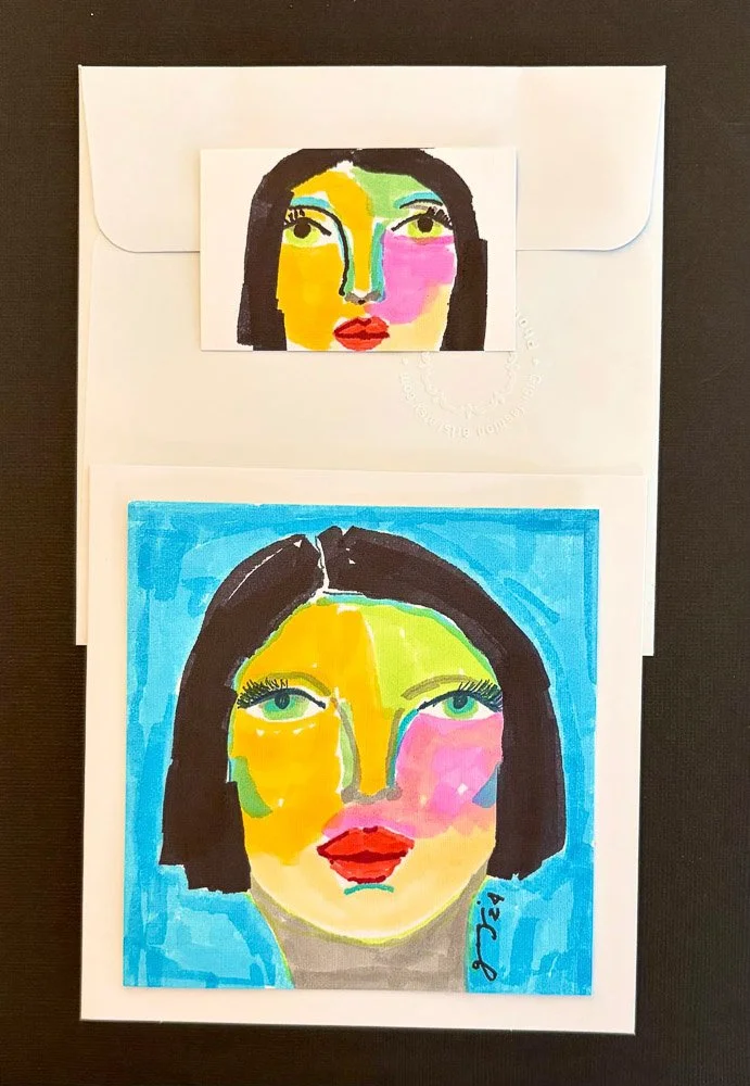 Colorful portrait of a woman with short black hair, green eyes, red lips, and lips with pink, yellow, and green splashes, painted on paper and placed on a white envelope.