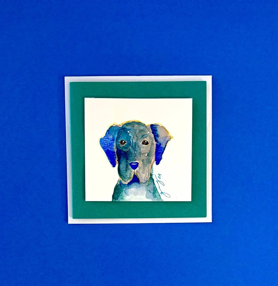 Watercolor painting of a dog's face with floppy ears, set against a white background and framed with a green mat, mounted on a blue wall.