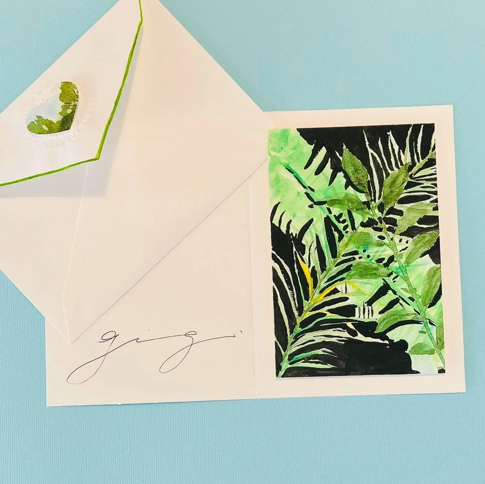 A greeting card with a botanical watercolor painting of green and black leaves, alongside an envelope with a green interior and a sticker of leafy greenery, set on a light blue surface.