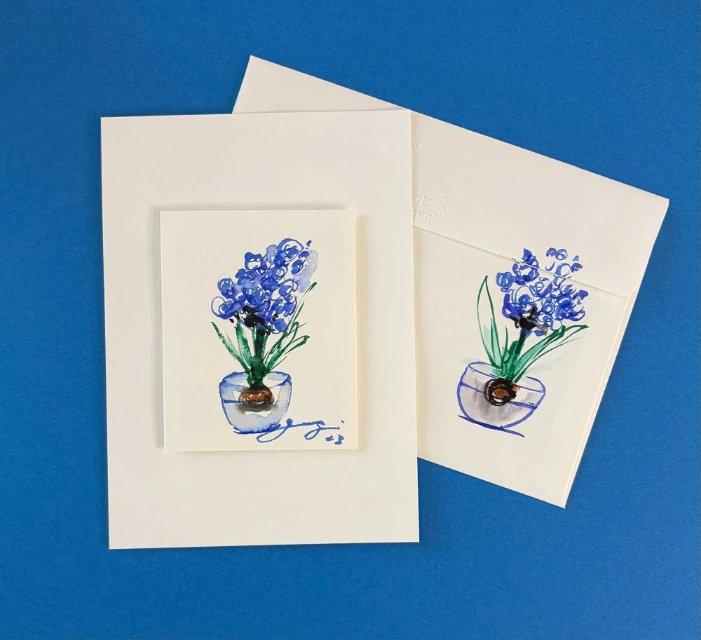 Two greeting cards with watercolor flower illustrations of blue flowers in a vase, one card is on a white envelope and the other is on a blue background.