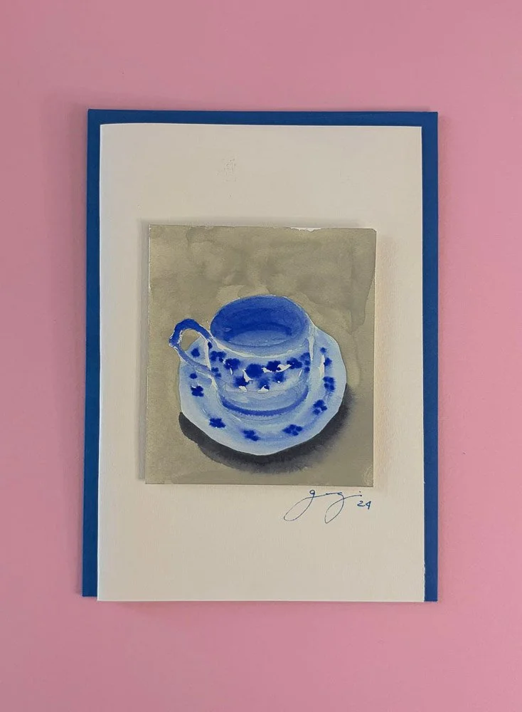 A watercolor painting of a blue and white patterned teacup and saucer on a gray background, signed 'g g' with the year 2021, mounted on white paper and framed with a blue border, placed on a pink wall.