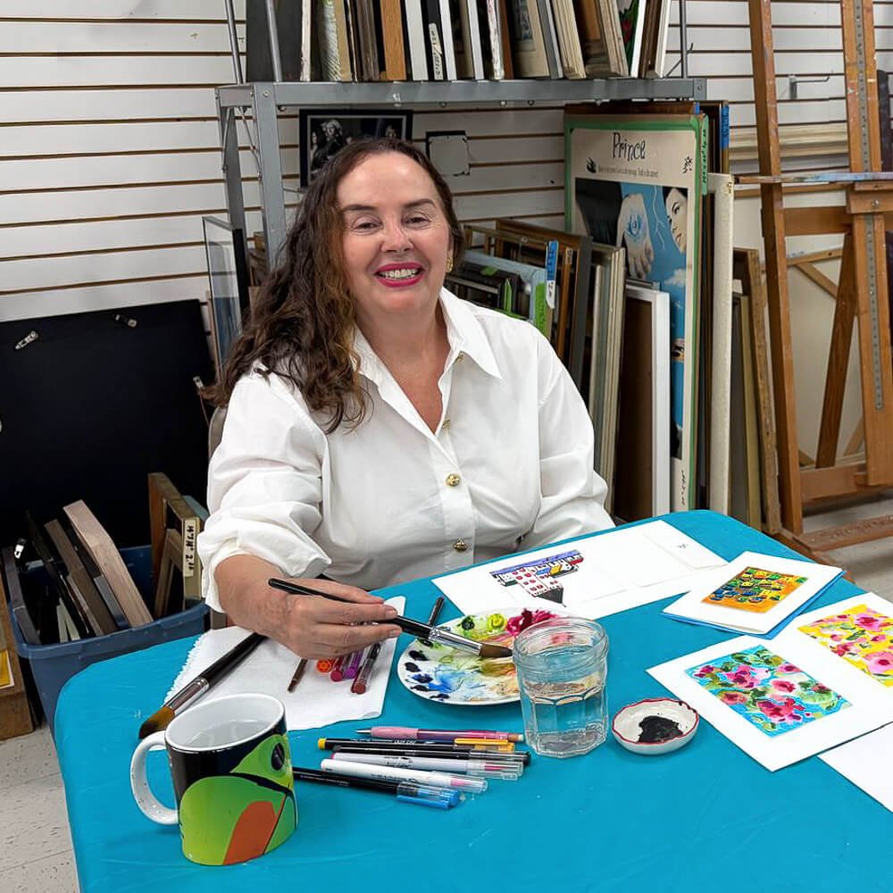 A woman with long curly hair and a white shirt sitting at a table with art supplies, smiling. There are colorful paintings, markers, a mug, and a glass of water on the table. Shelves with books and framed photographs are in the background.