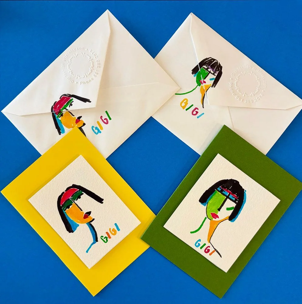 Two handmade cards with colorful abstract portraits of women and the word 'GIGI' printed beneath. The cards are on a blue surface, one with a yellow border and the other with a green border. Two envelopes with matching artistic portraits and 'GIGI' are also present.