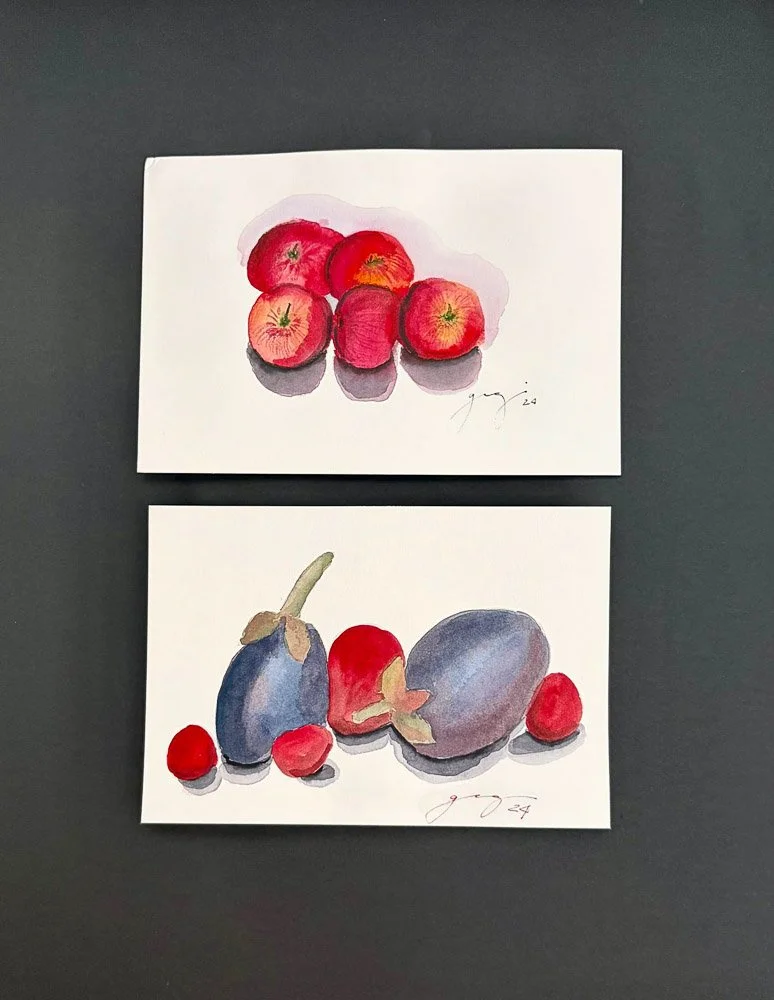 Two watercolor paintings of apples and berries on white paper placed on a dark surface. The top painting depicts red apples, while the bottom painting features purple eggplants and red berries.