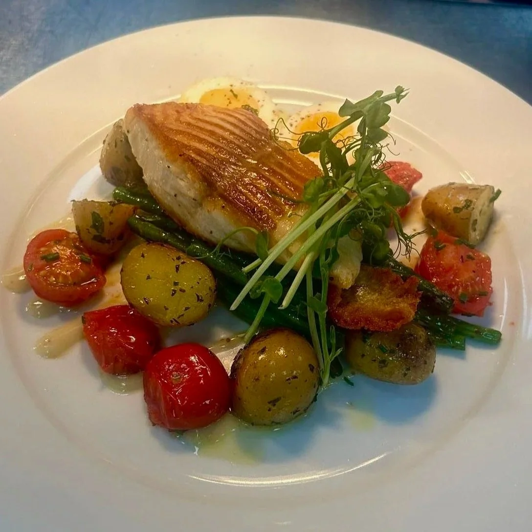A plated dish featuring a grilled piece of fish, cherry tomatoes, potatoes, and greens, garnished with microgreens.
