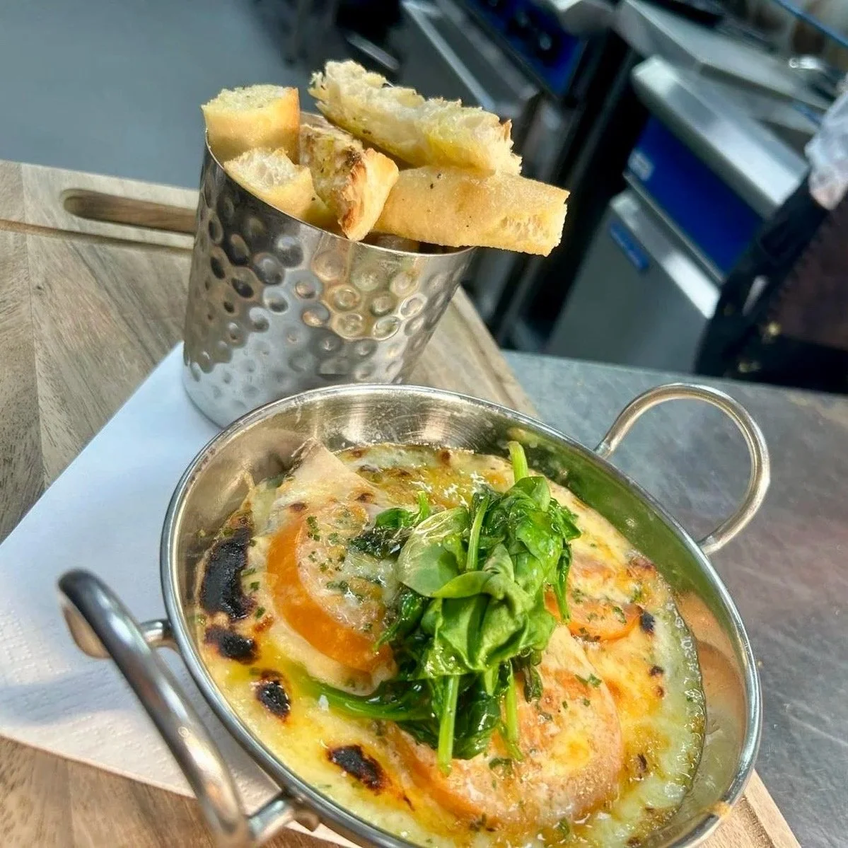Baked eggs in a small metal dish topped with greens, served with a side of bread in a metal container.