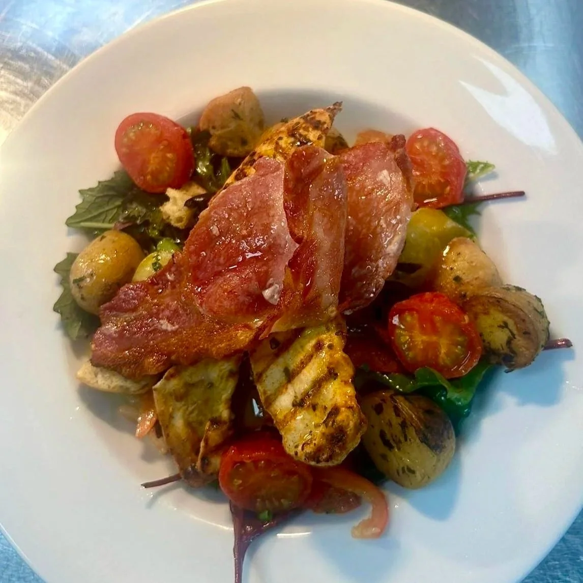 Grilled chicken dish with cherry tomatoes, potatoes, lettuce, and bacon on a white plate.