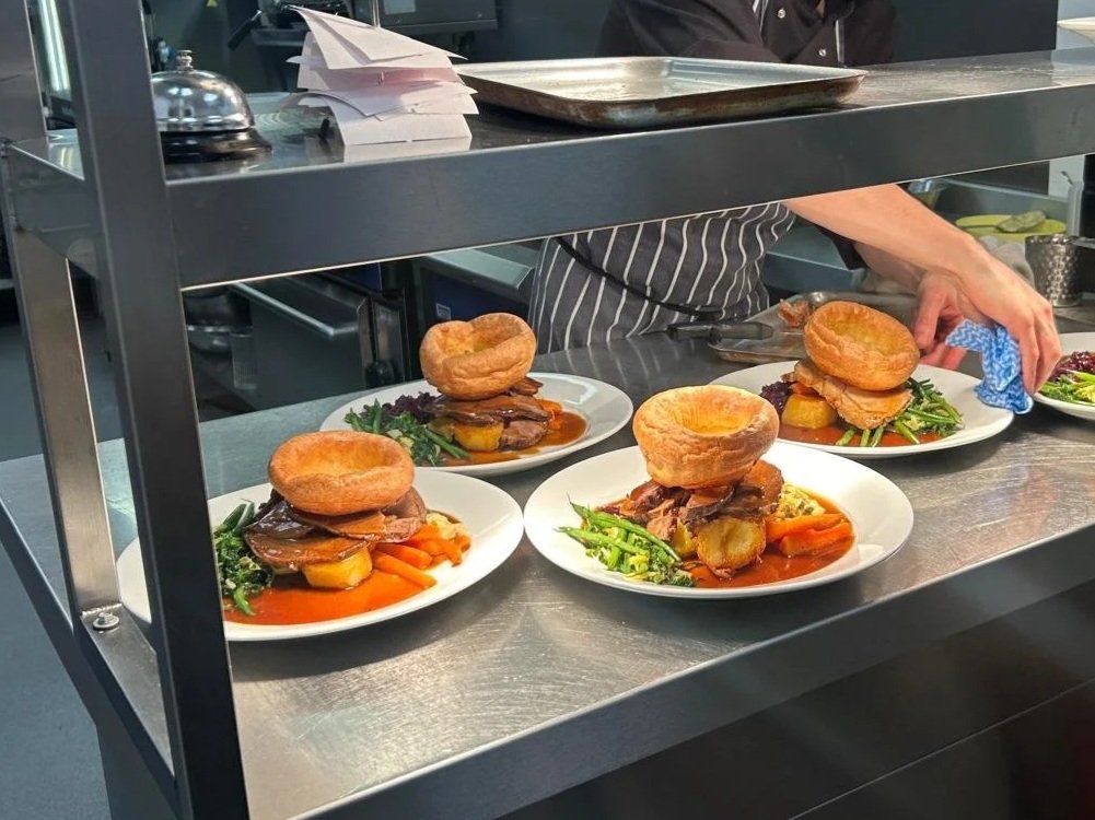Three plates of food on a kitchen counter, each containing a large Yorkshire pudding, roast meat, vegetables, and gravy, with a person in a striped apron preparing the dishes.