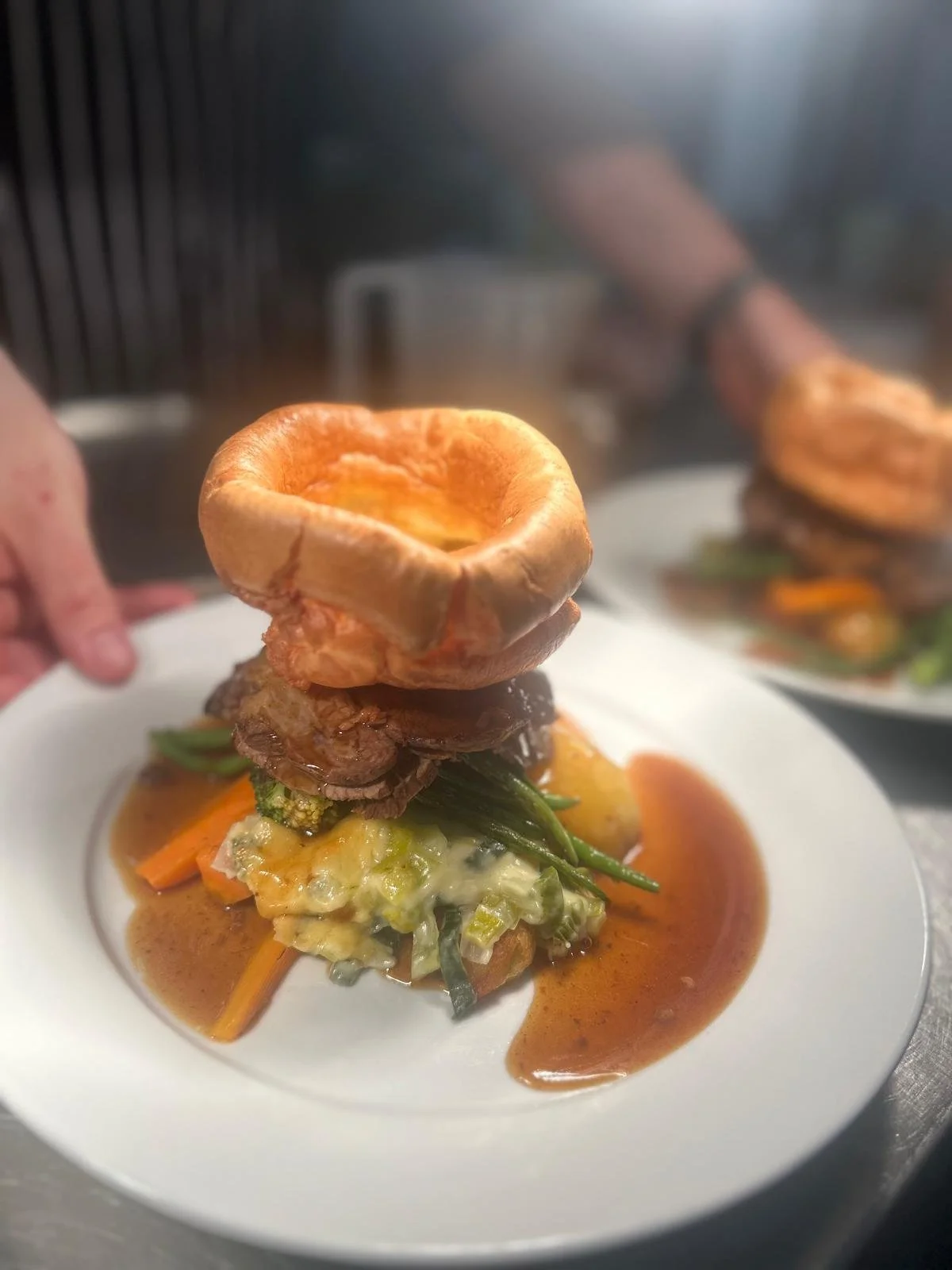 A gourmet dish with a baked shell filled with sauce on a bed of vegetables and mashed potatoes, topped with a meat patty and a baked bread bowl.