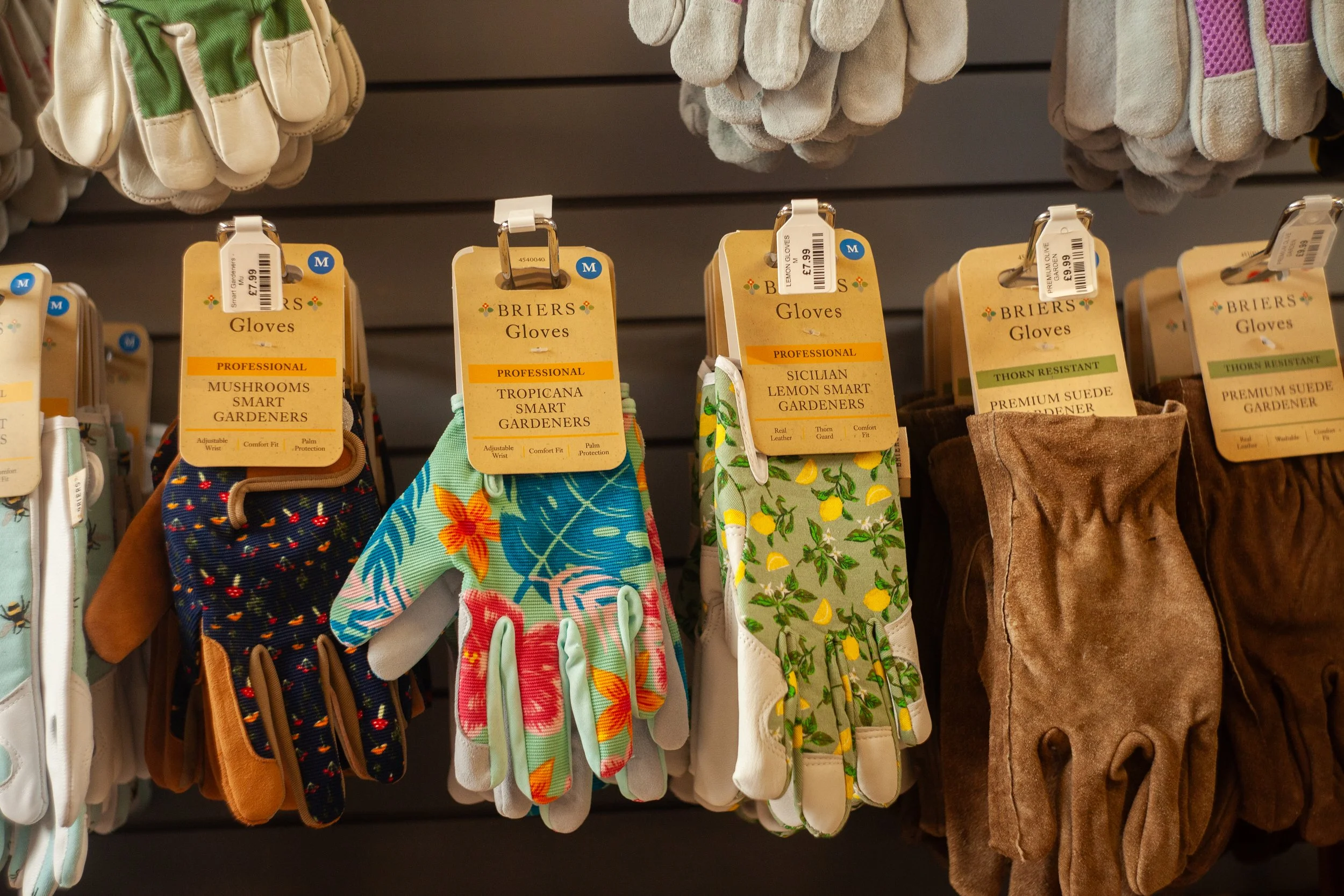 Display of gardening gloves with colorful floral and fruit patterns and text labels indicating professional, smart gardeners, and thistle resistance.