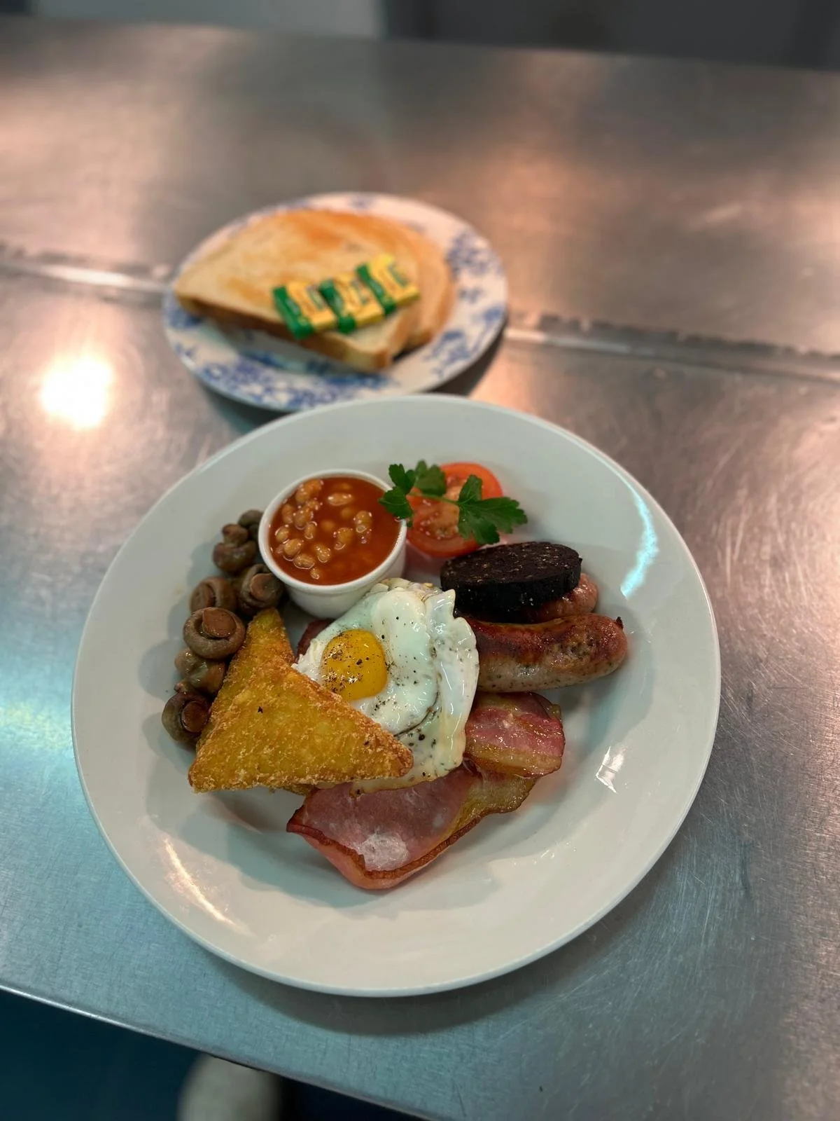 Plate of cooked breakfast with baked beans, fried egg, ham, sausage, black pudding, grilled tomato, and hash brown with a side of buttered toast on a floral plate in the background.