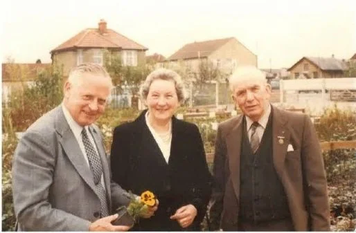 Three older people smiling outdoors, with a man holding a small bouquet of flowers, against a background of houses and a garden.