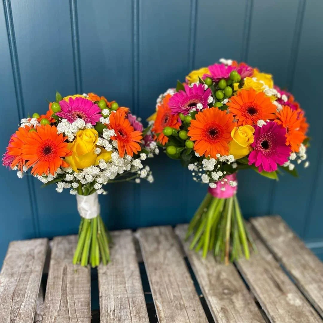 Two colorful bouquets of flowers with pink, orange, yellow, and white blooms, arranged on a rustic wooden surface against a blue paneled wall.