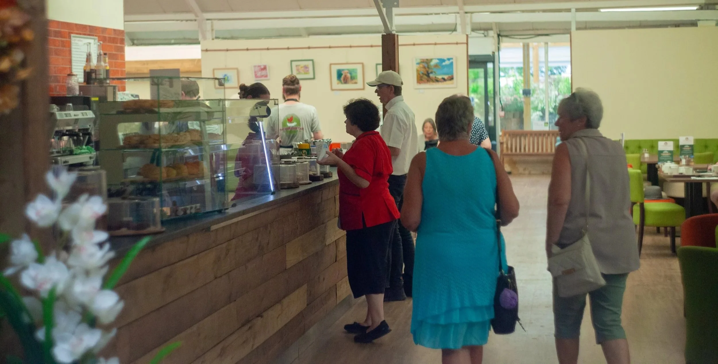 People standing in line at a cafe counter inside a restaurant with colorful framed artwork on the wall.
