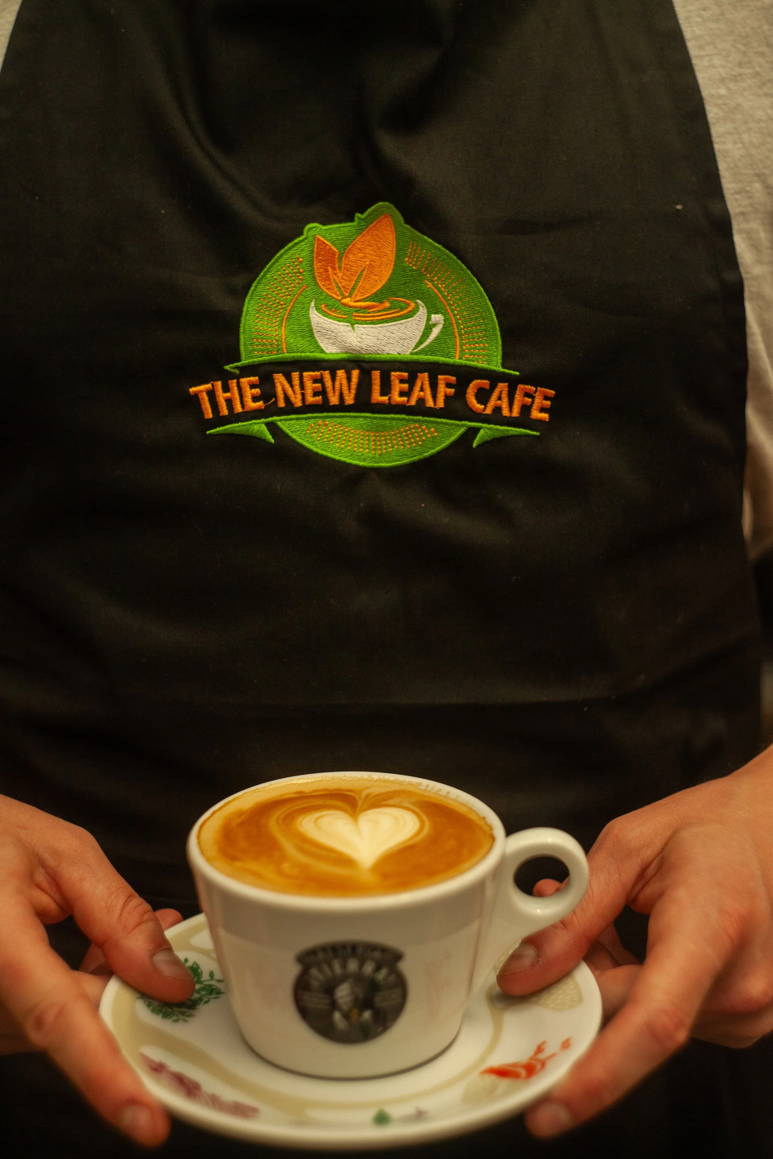 A person wearing a black apron with the logo and name 'The New Leaf Cafe' embroidered on it is holding a white ceramic cup of latte with a heart-shaped foam design on top. The cup is placed on a saucer with a decorative design.