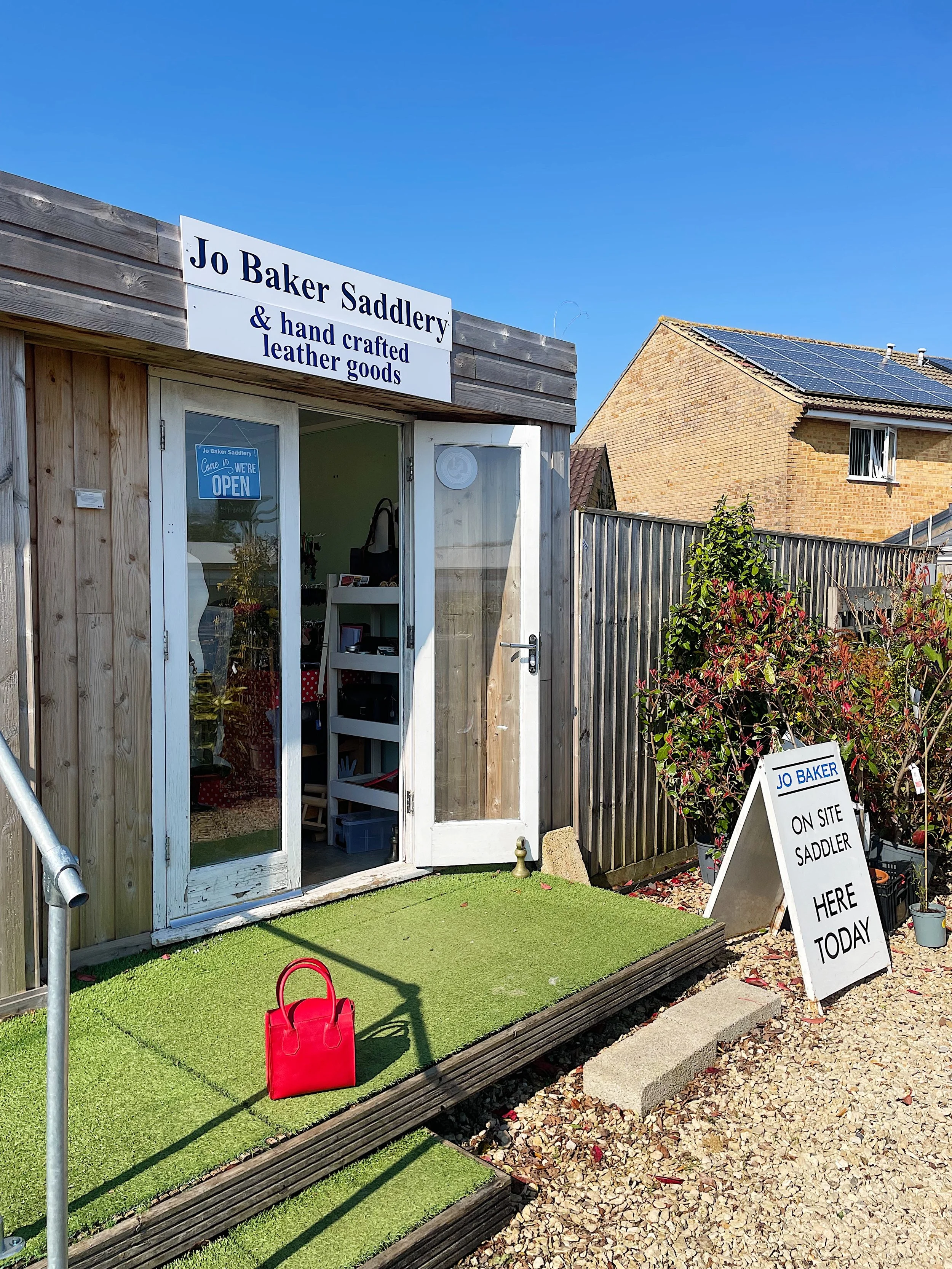 Small storefront with a sign that says 'Jo Baker Saddlery & hand crafted leather goods'. A glass door with a blue 'Come in, we're open' sign. A white sandwich board sign on the gravel reads 'Jo Baker Saddler on site, here today'. A red handbag is on the green artificial turf outside. A building with solar panels is visible in the background under a clear blue sky.