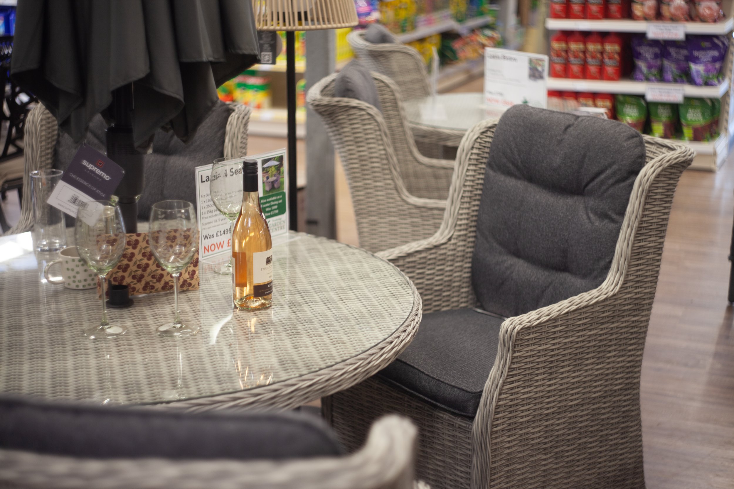 A patio dining setup inside a retail store featuring wicker chairs with gray cushions, a glass table with wine glasses, a bottle of rosé wine, and various signs and price tags.
