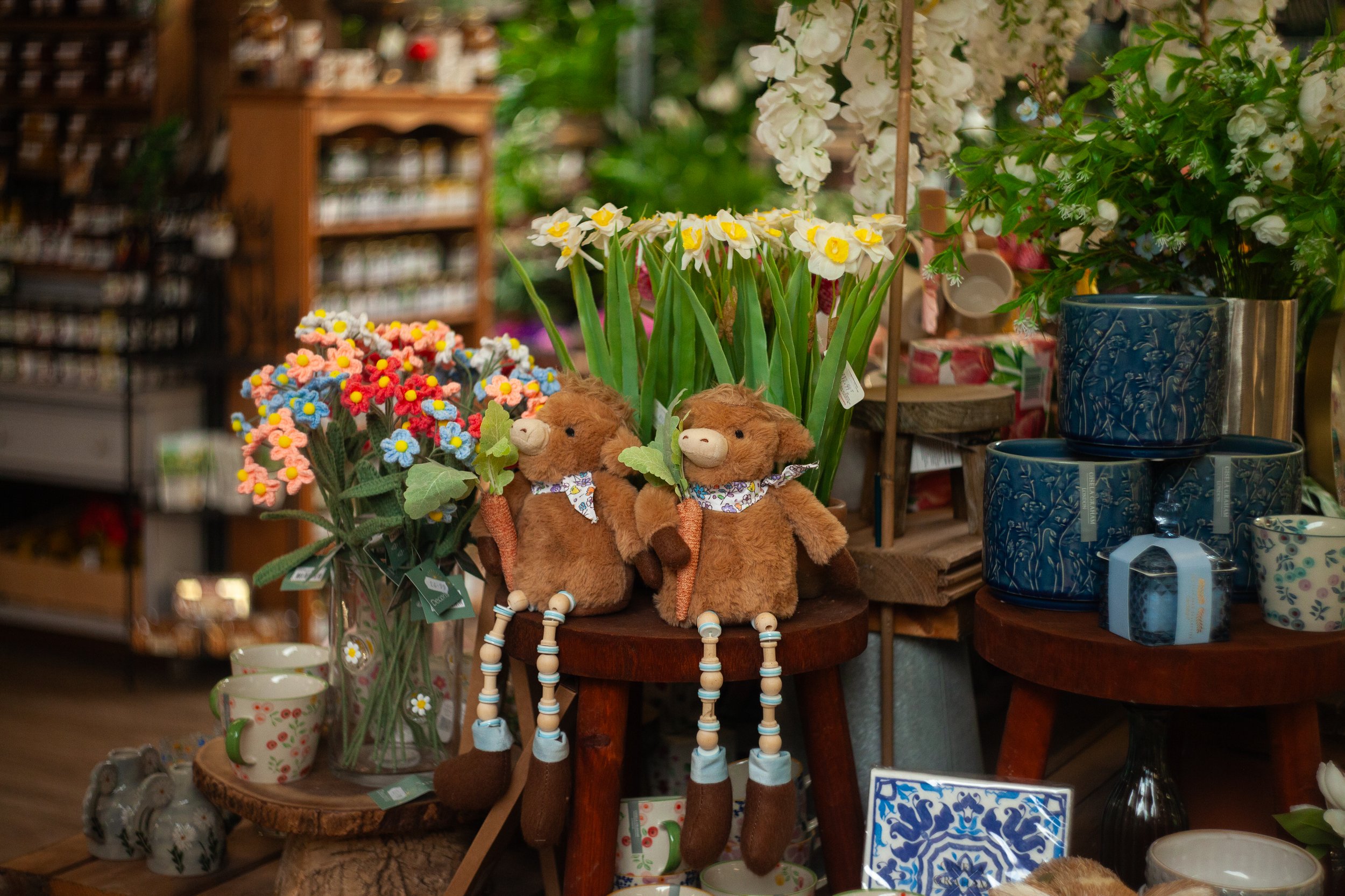A display of colorful artificial flowers, plush toys resembling llamas, wooden beaded jewelry, ceramic mugs, and decorated pots in a store or market setting.