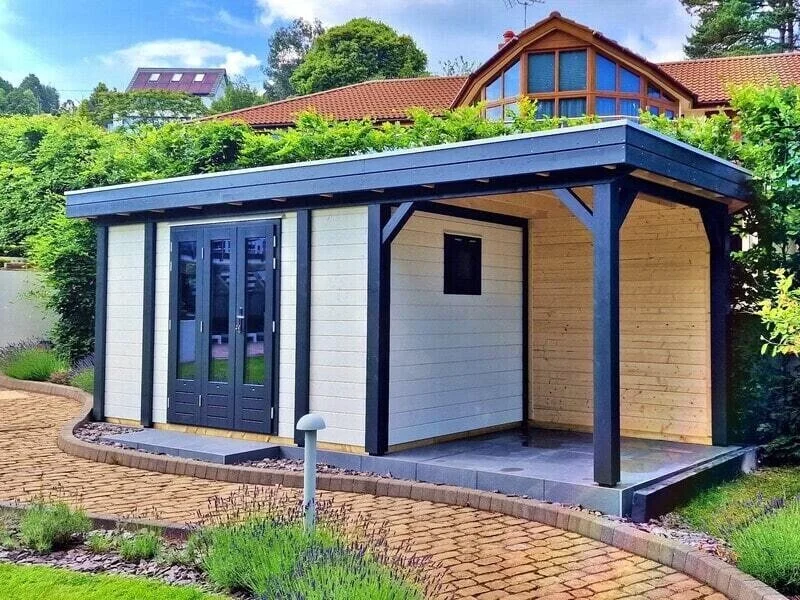 A small modern shed with white siding, black trim, and an attached covered porch, situated in a garden with a brick pathway. There are trees and a house in the background.