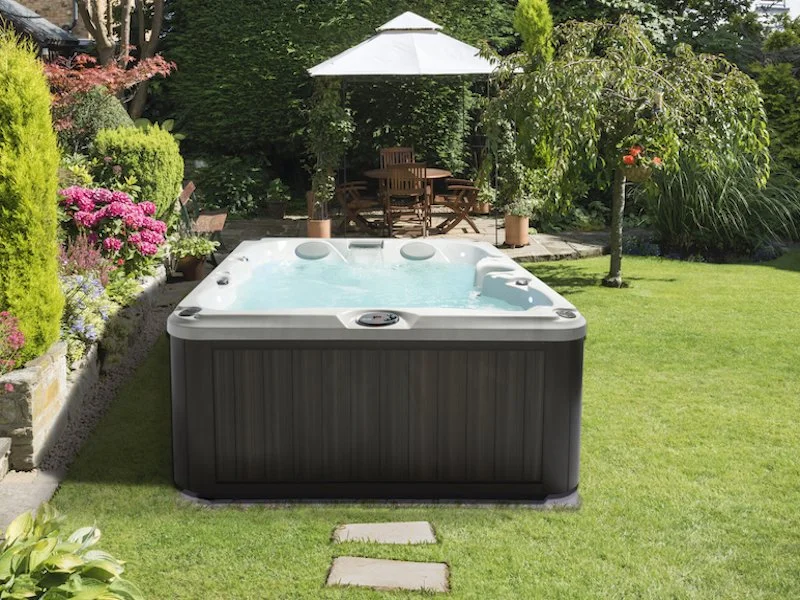 Hot tub in a lush backyard garden with a wooden patio and table, surrounded by greenery, flowers, and a tree.
