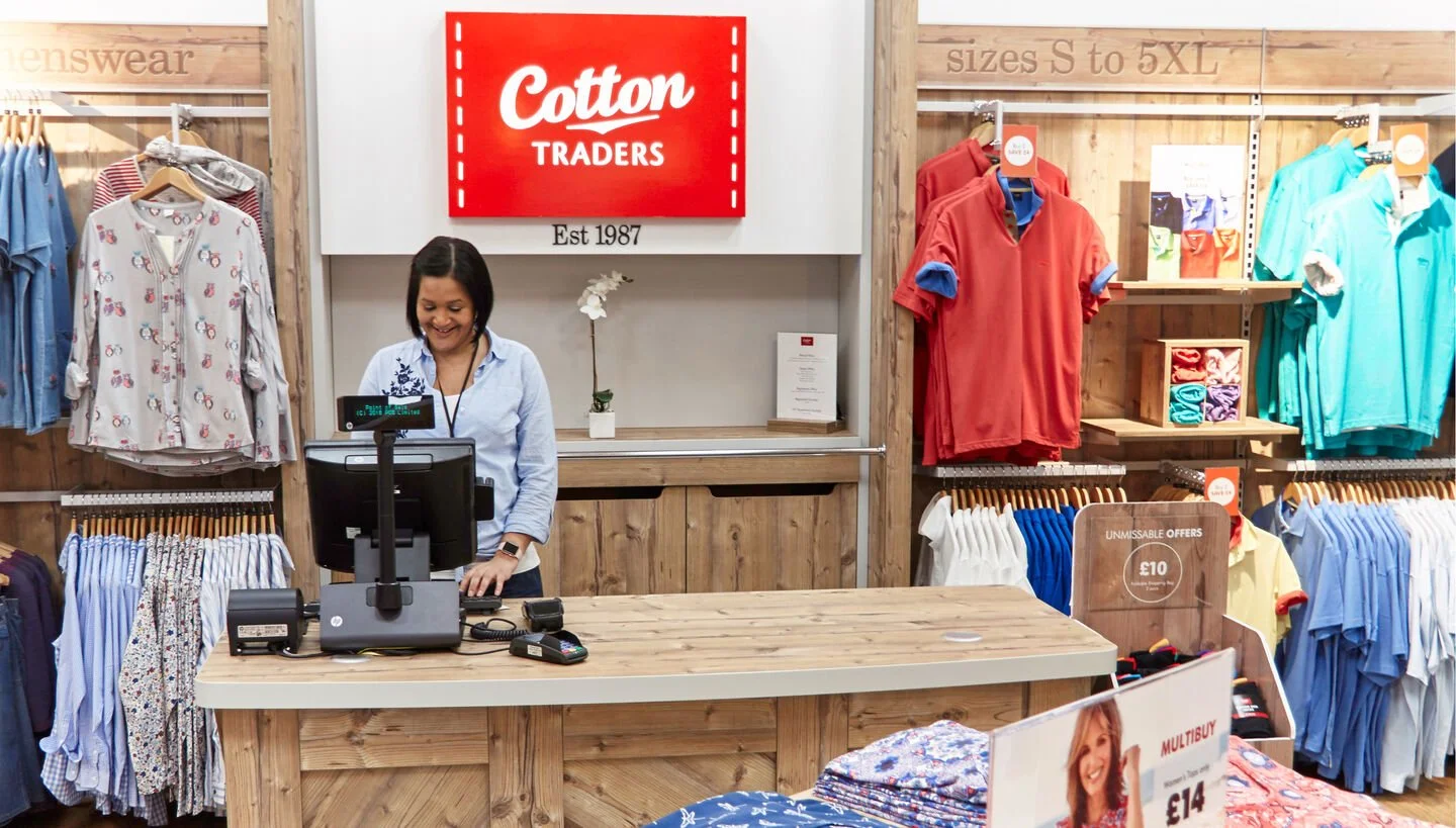 Inside a clothing store, a smiling woman checks out at the cashier desk. Racks of colorful shirts and blouses are on the walls around her, with signage indicating sizes and special offers.