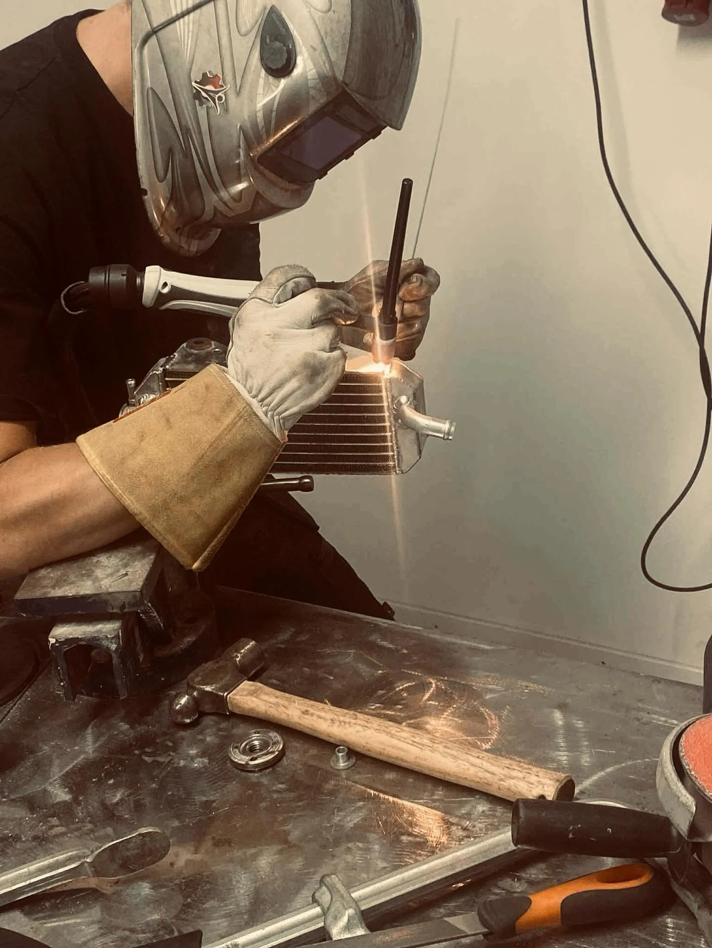 En person svetsar en metallkylare med ett svetsverktyg i ett verkstadshörn. Personens händer bär krats- och svetsutrustning. Verktyg, metalldelar och arbetsplatsen syns på arbetsbänken.