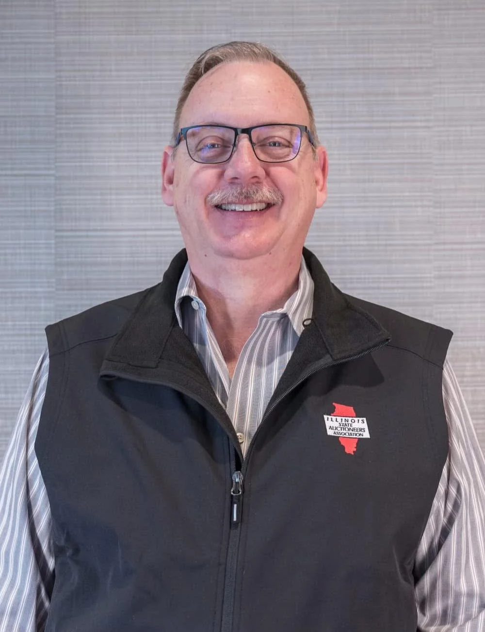 A smiling middle-aged man with glasses and a mustache, wearing a black vest with an Illinois State Auctioneers Association logo, and a striped button-up shirt, standing against a gray textured wall.