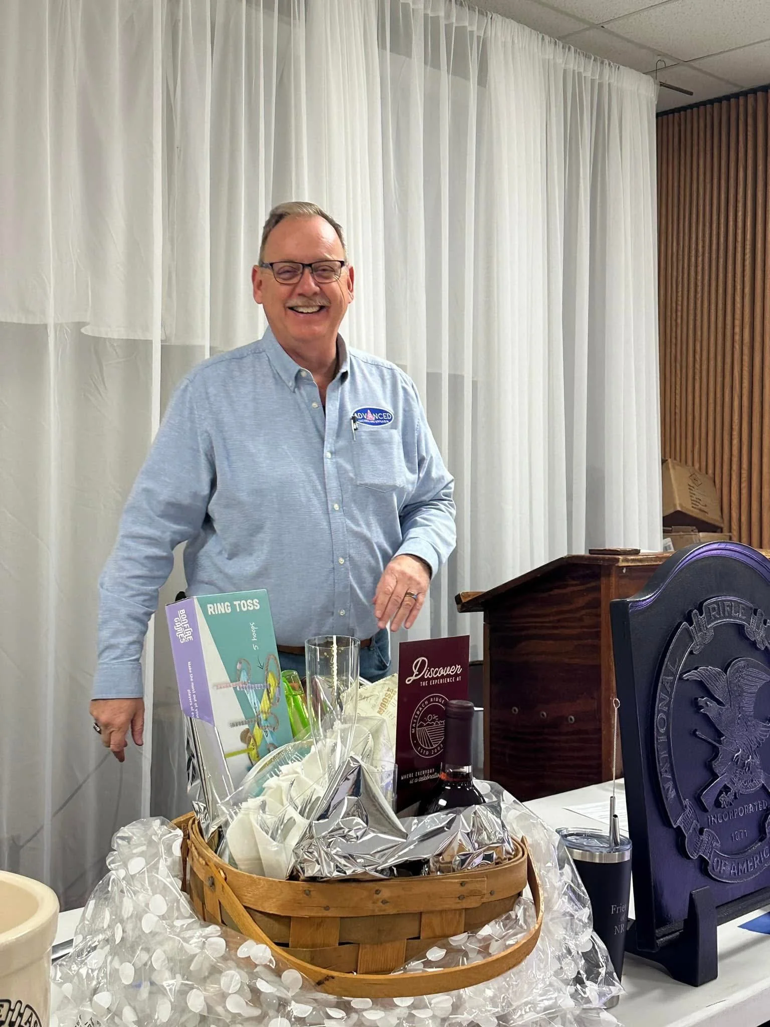 Man in glasses and light blue button-down shirt smiling at a table with wrapped gifts, a wine bottle, and promotional signs.