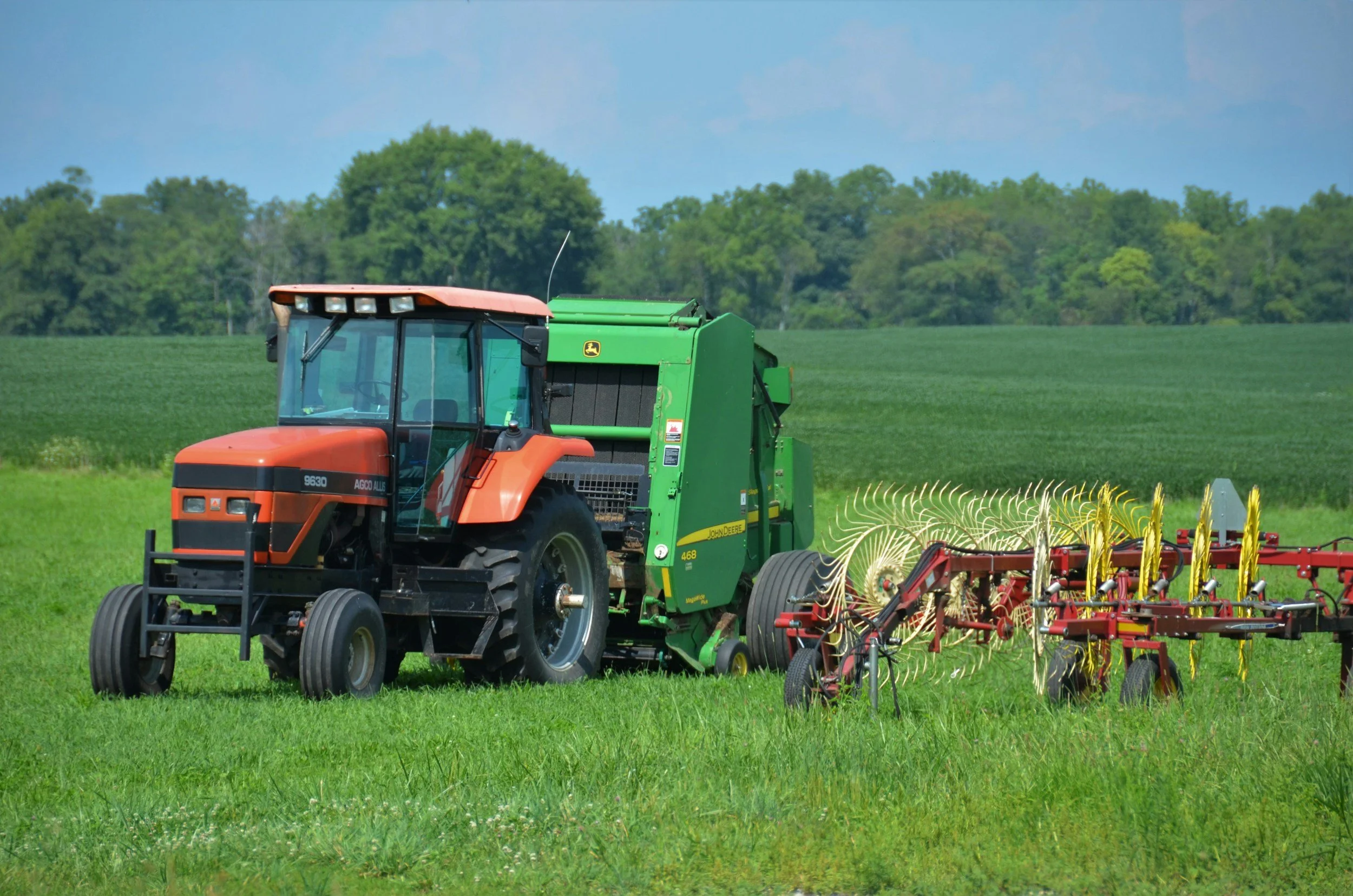 A tractor pulling a hay rake in a green field, with trees and a cloudy sky in the background.