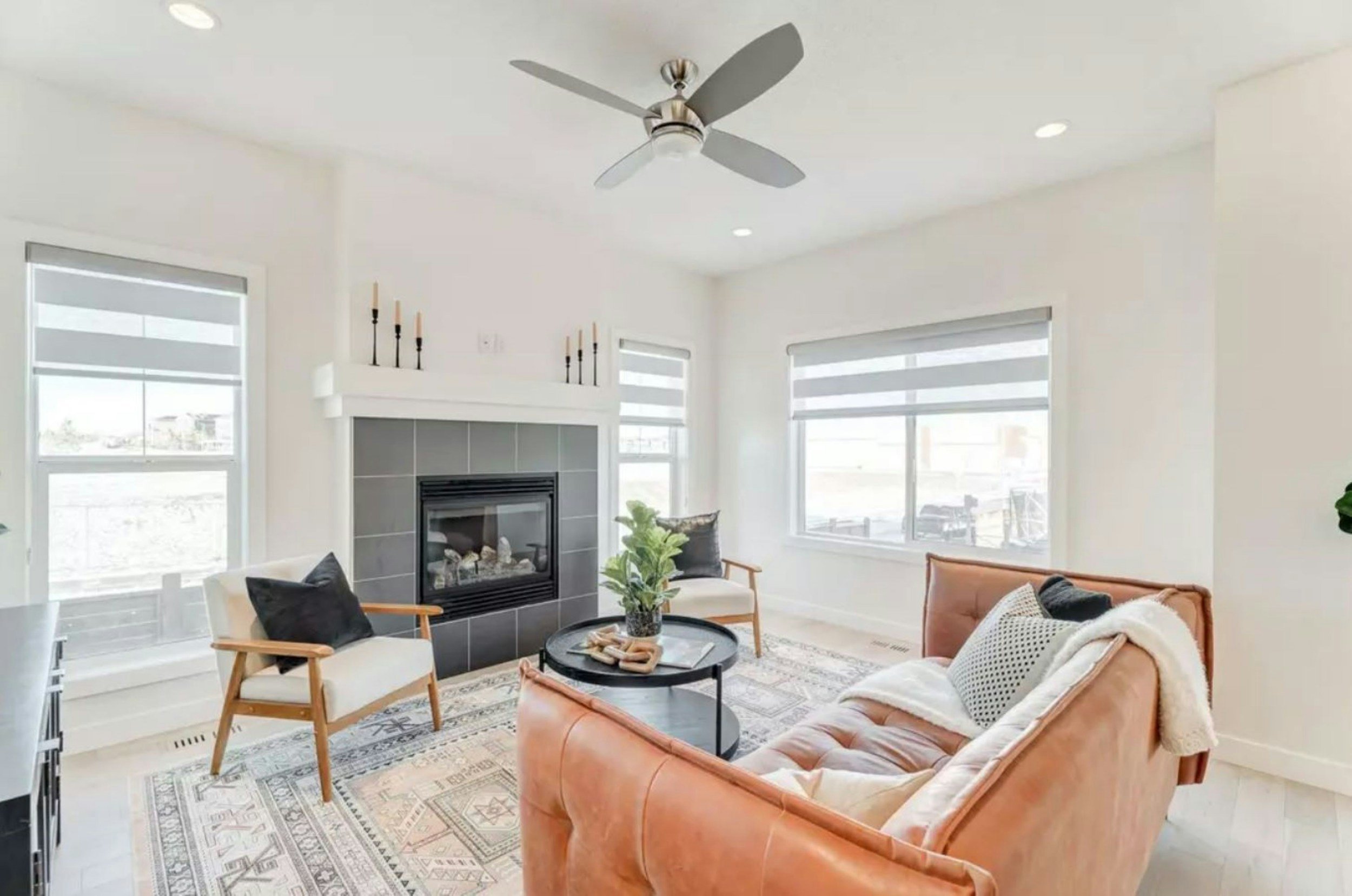Bright living room with white walls, a ceiling fan, large windows with horizontal blinds, a fireplace with gray tiles, a tan sofa with pillows, two armchairs, a black round coffee table with decorative items, and a patterned rug.