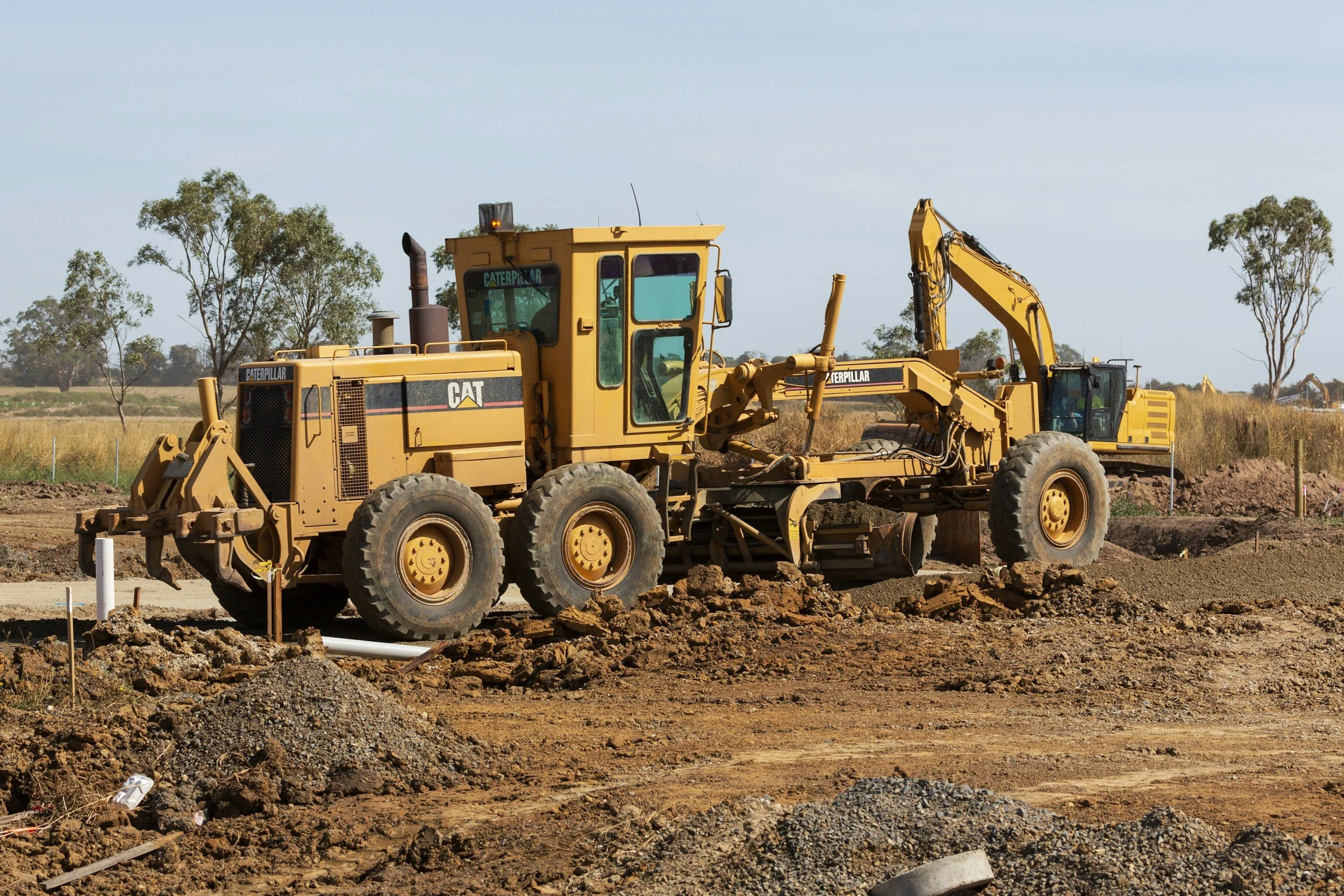 A yellow Caterpillar bulldozer working on a construction site with dirt, rocks, and scattered construction materials, and a background of trees under a blue sky.