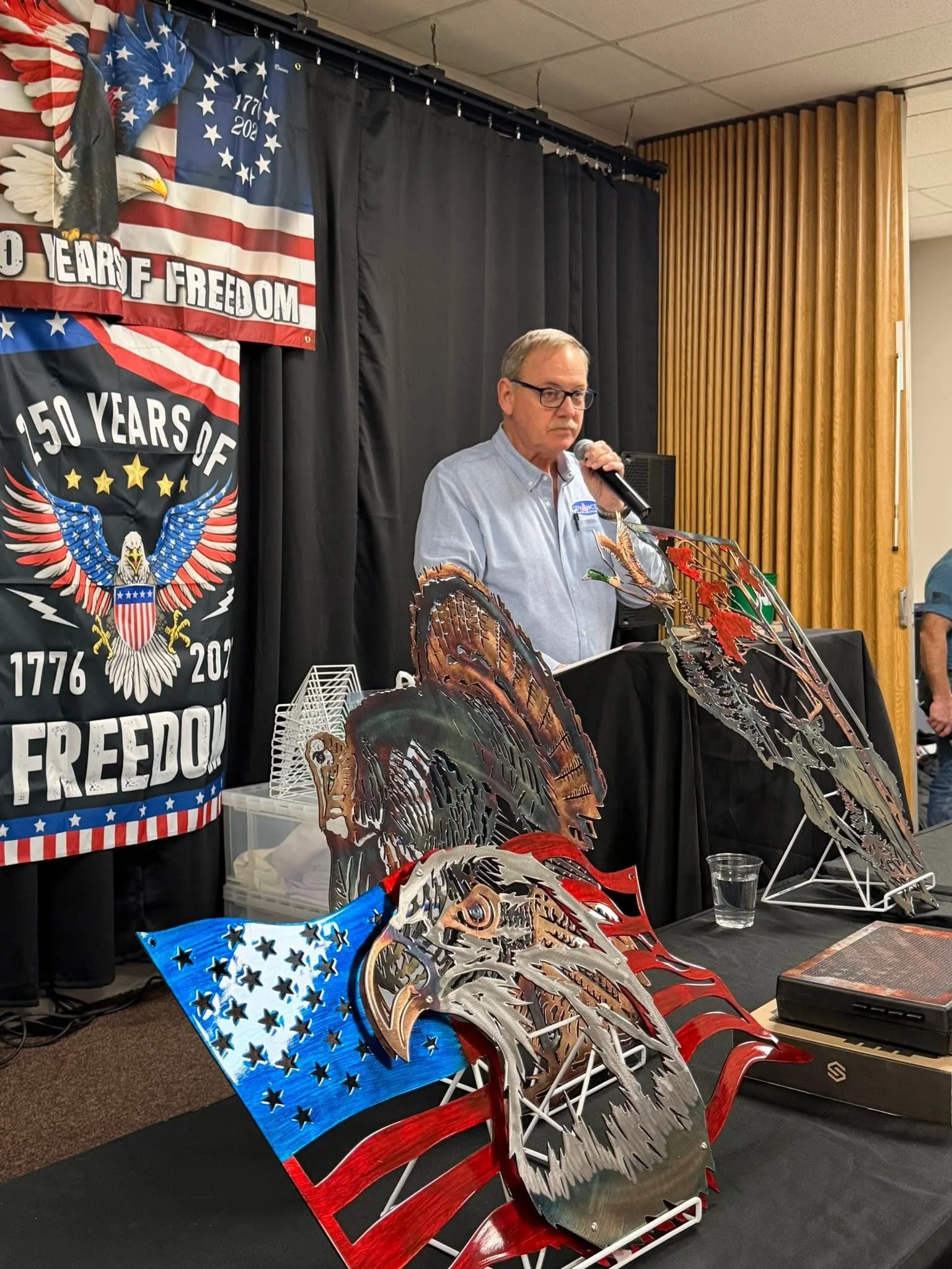 A man with glasses speaking into a microphone at a table with metal eagle sculptures and an American flag-themed design, with banners in the background celebrating 150 years of freedom and features of an eagle with the American flag.