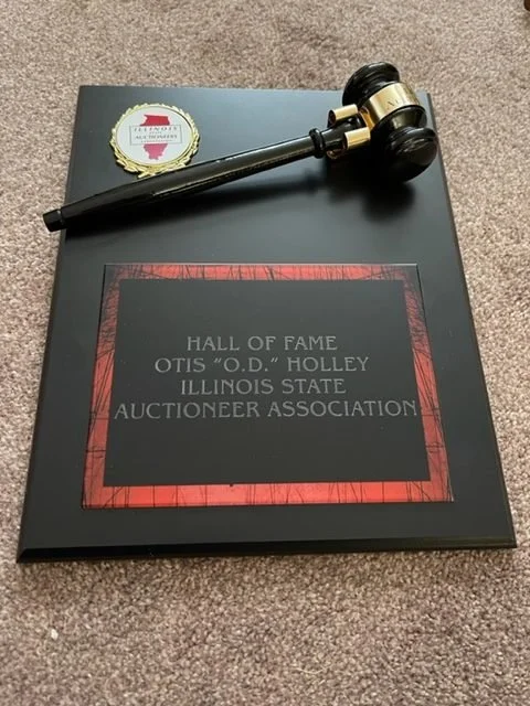 Gavel resting on a plaque that reads 'Hall of Fame Otis "O.D." Holley Illinois State Auctioneer Association,' with a sticker in the top left corner.