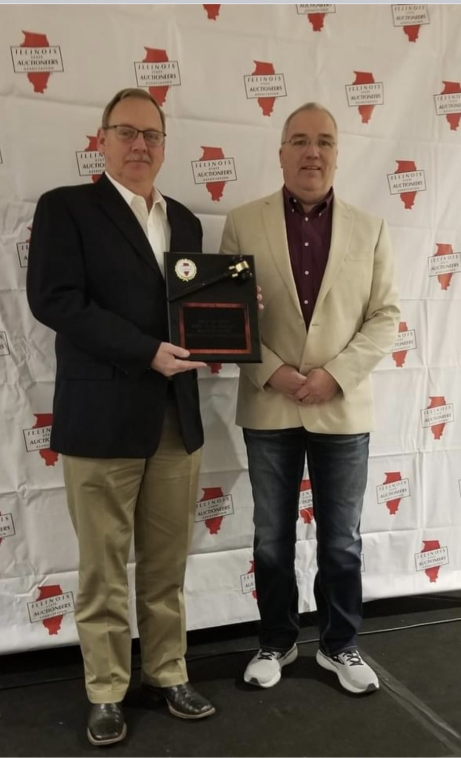 Two men standing in front of a backdrop with the Illinois auctions logo. One man is holding a plaque and a gavel, both smiling.