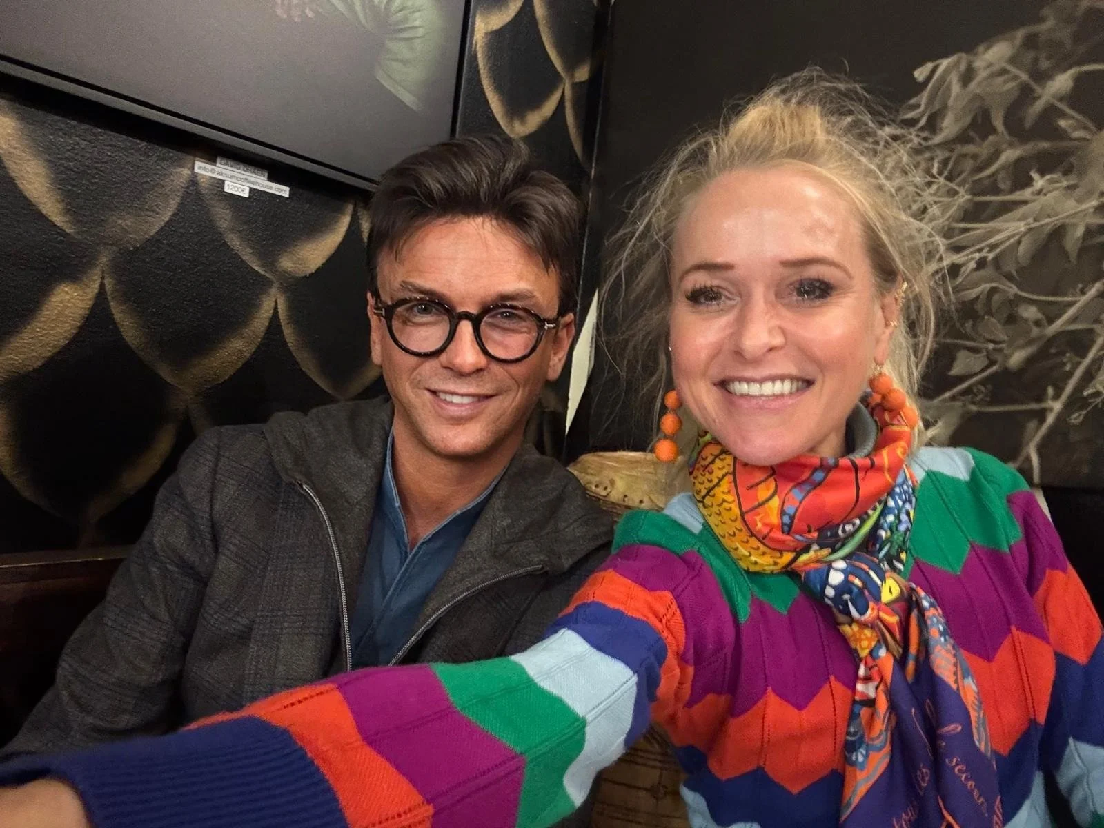 A man and woman smiling and taking a selfie together in front of a dark background with abstract patterns.