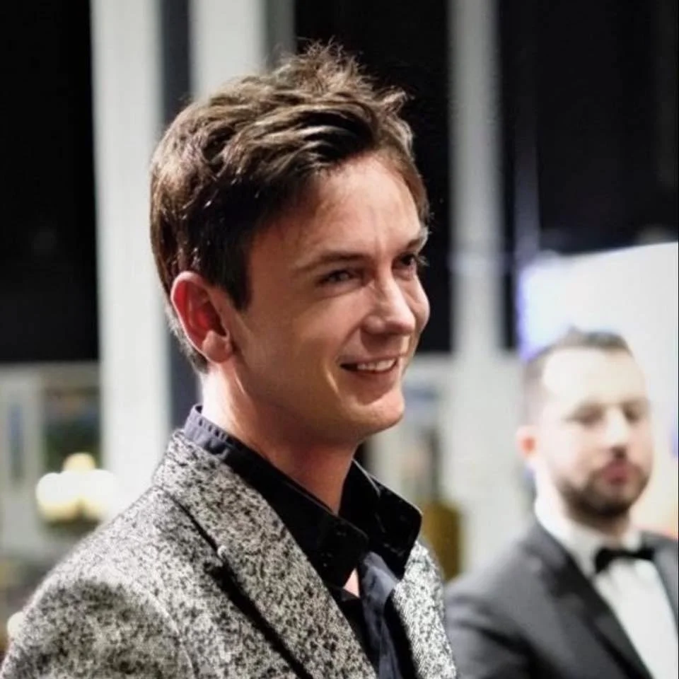 Young man smiling, wearing a suit with a patterned blazer and a black shirt, at a formal event.