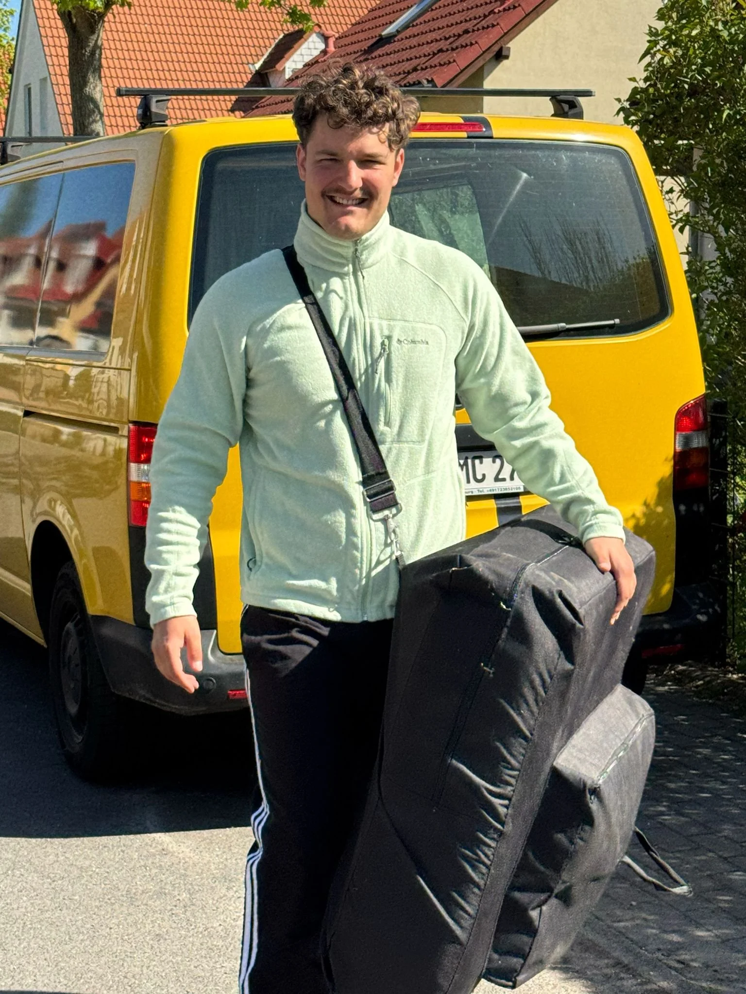 Ein lachender Mann mit braunen, lockigen Haaren trägt eine weiße Fleece-Jacke und eine schwarze Tasche über der Schulter. Er steht neben einem schwarzen Rucksack, während er in Richtung eines gelben Lieferwagens geht, der im Hintergrund geparkt ist. Es ist ein sonniger Tag, und die Umgebung scheint ein Wohngebiet zu sein.