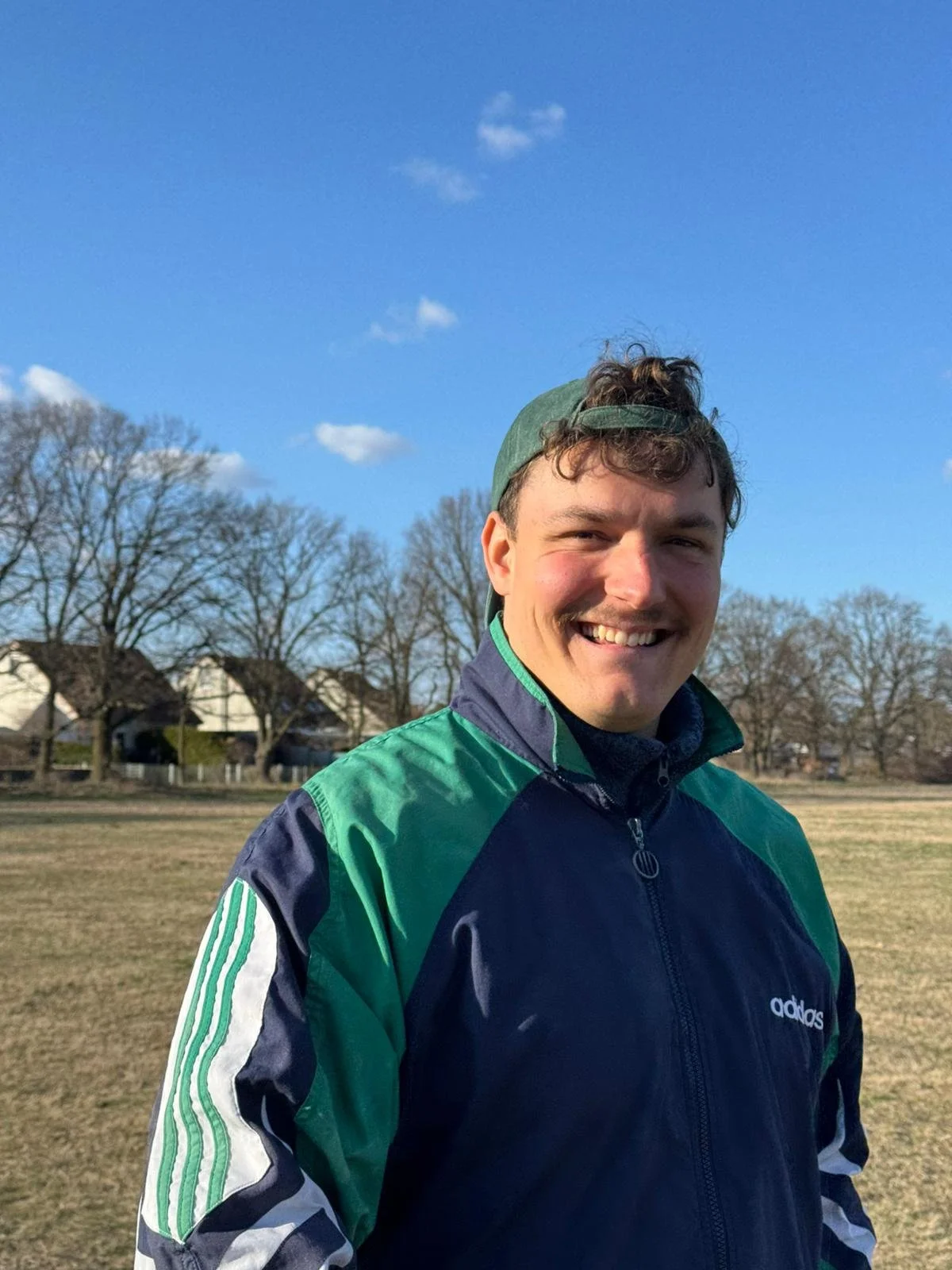 Lächelnder junger Mann in einer grünen und blauen Adidas-Jacke im Freien bei Sonnenschein, mit Bäumen und Häuser im Hintergrund.