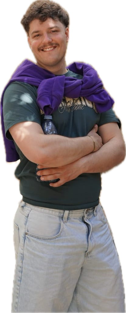 Ganzköperfoto von Lukas. Lächeln, verschränkte Arme. Ein lila Pullover liegt an den Ärmeln verknotet auf den Schultern. Er trägt eine hellblaue Jeans und ein grünes T-Shirt.