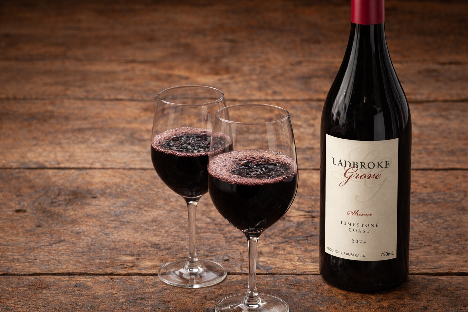 Two glasses of red wine and a bottle of Ladbroke Grove Shiraz Limstone Coast 2024 on a wooden surface.