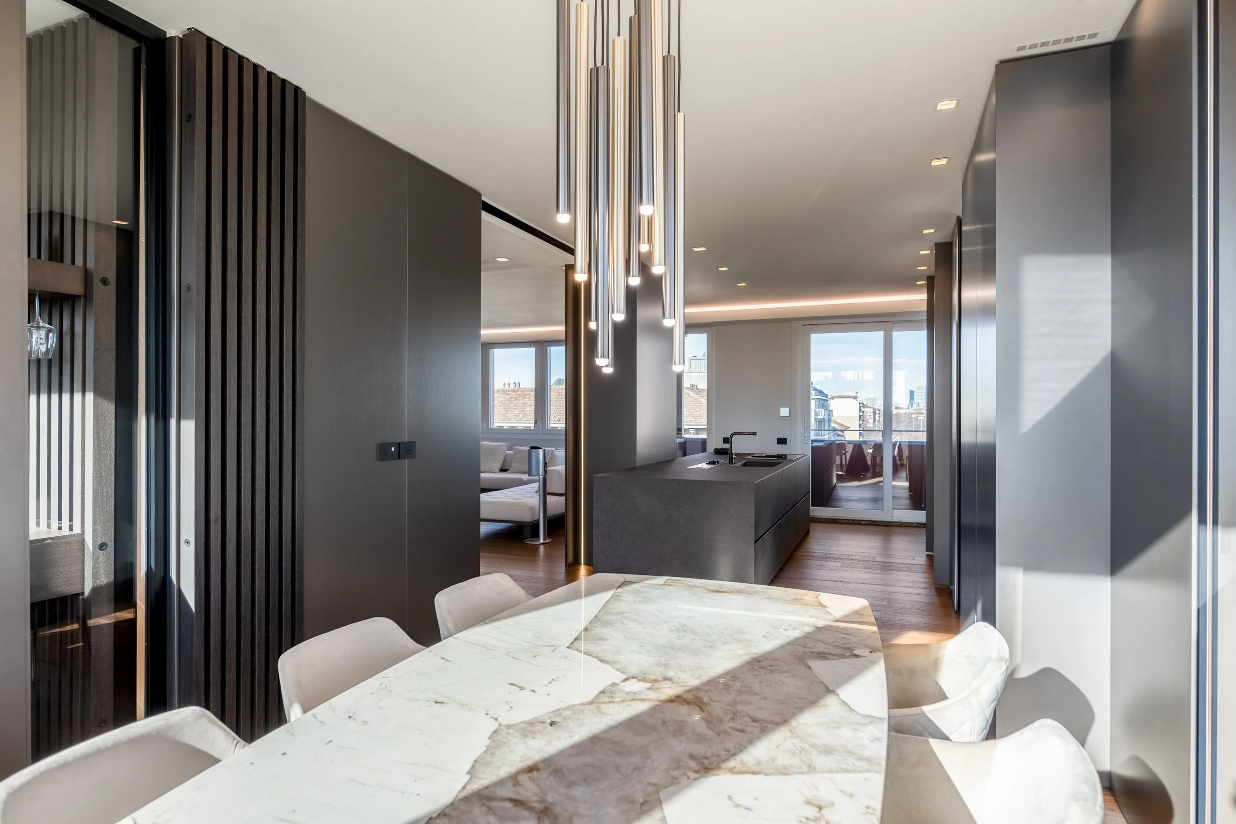 Interno moderno con tavolo in marmo, sedie bianche, cucina con isola nera, illuminazione a sospensione lucida, finestre grandi con vista sulla città