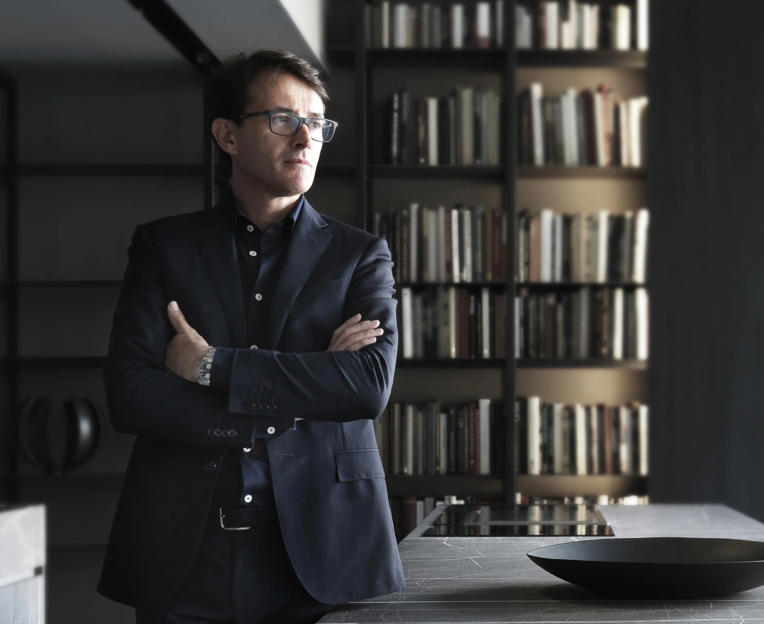Un uomo in abito nero con occhiali da vista si trova in un ambiente moderno con scaffali pieni di libri sullo sfondo, e un tavolo di piastrelle grigie con una grande ciotola nera in primo piano.