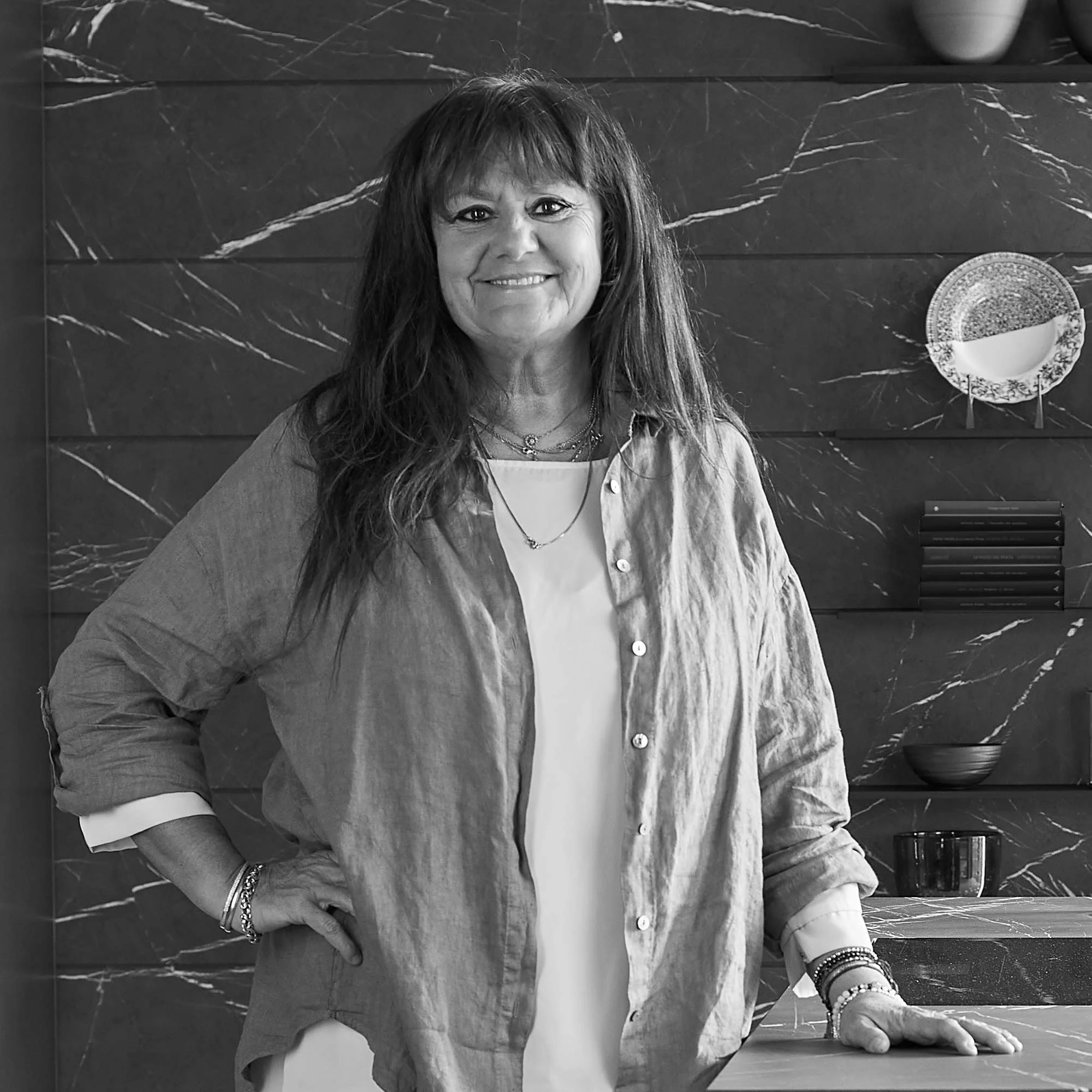 Donna sorridente con capello lungo e cappotto leggero davanti a uno sfondo di lastre di marmo scuro con decorazioni di ceramica e libri.