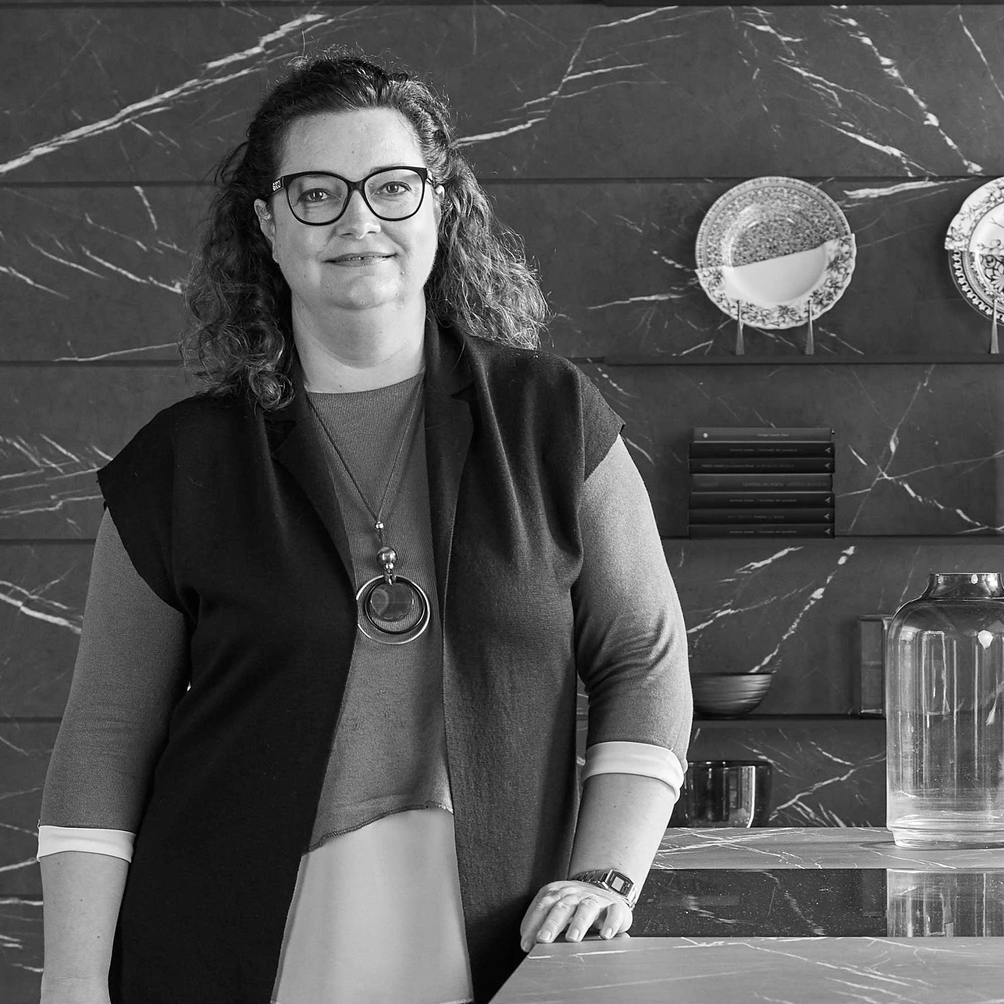 Una donna sorridente con occhiali, indossa una maglietta a maniche lunghe e un gilet e si trova in una cucina moderna con pareti in marmo scuro e piatti decorativi appesi.